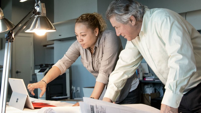 Zwei Mitarbeiter, eine Frau und ein Mann, im Gespr&auml;ch an einem Tisch mit Architektur-Pl&auml;nen und einem Laptop