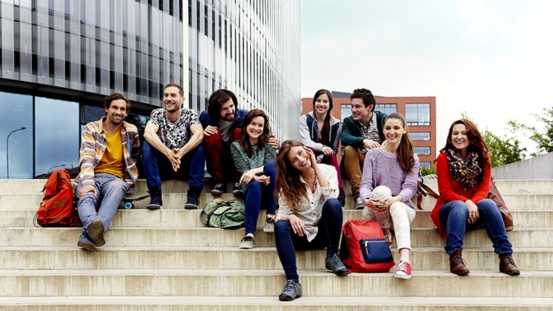 Mehrere Studentinnen und Studenten sitzen auf einer Treppe vor einem Geb&auml;ude