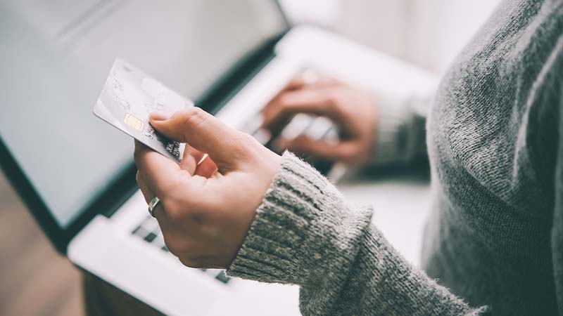 Eine Person bedient mit ihrer rechten Hand einen Laptop, w&auml;hrend ihre linke eine Bankkarte h&auml;lt