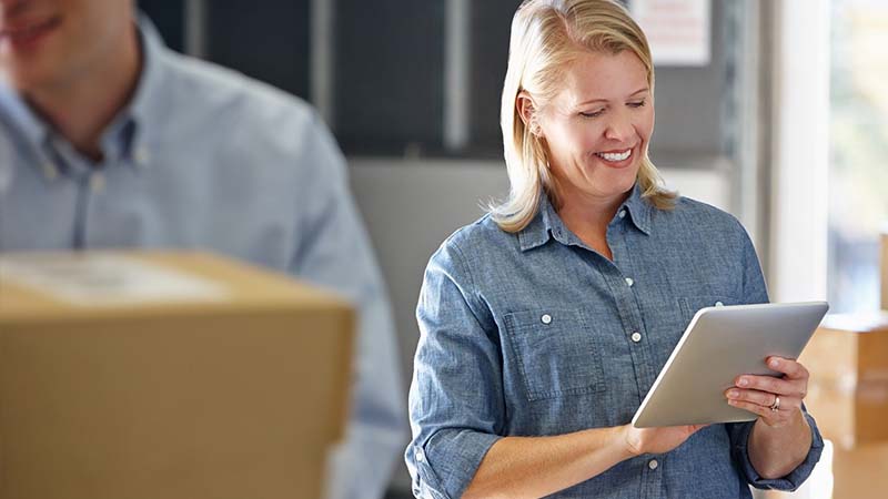 Eine Frau, umgeben von Paketen, blickt auf einen Tablet Computer