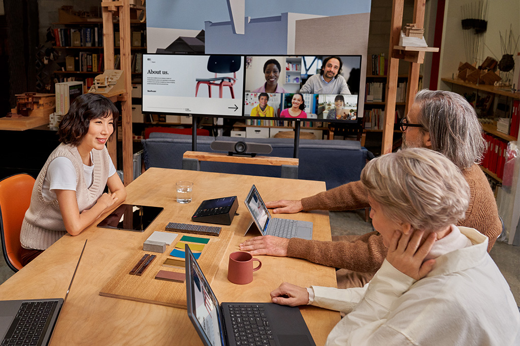 Participants sit at a table looking at a large screen during a Teams meeting