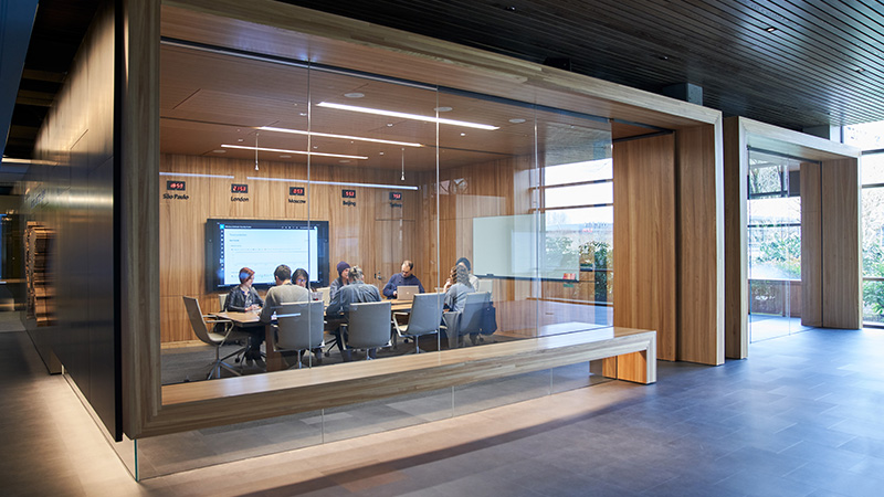 A group of employees sit around a table, working at their laptops during a meeting in a glass-walled office with large screen