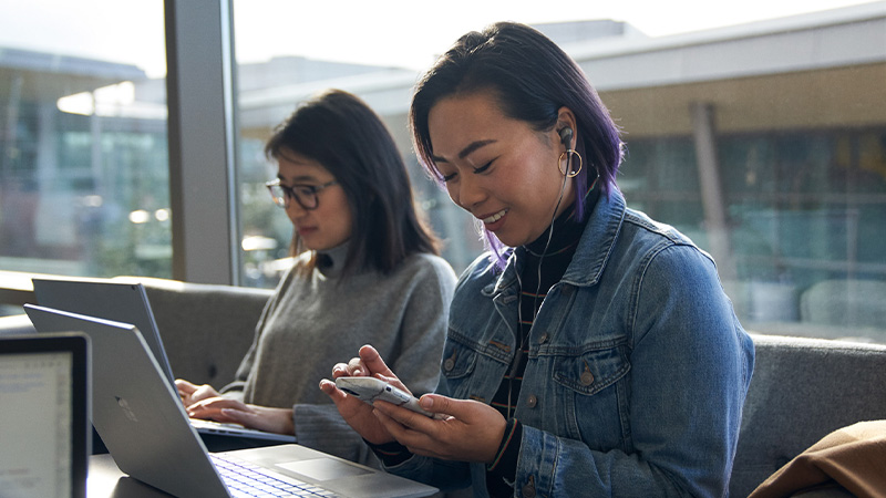 Two people sitting indoors by large windows, working on laptops, with one person also using a smartphone and wearing earbuds