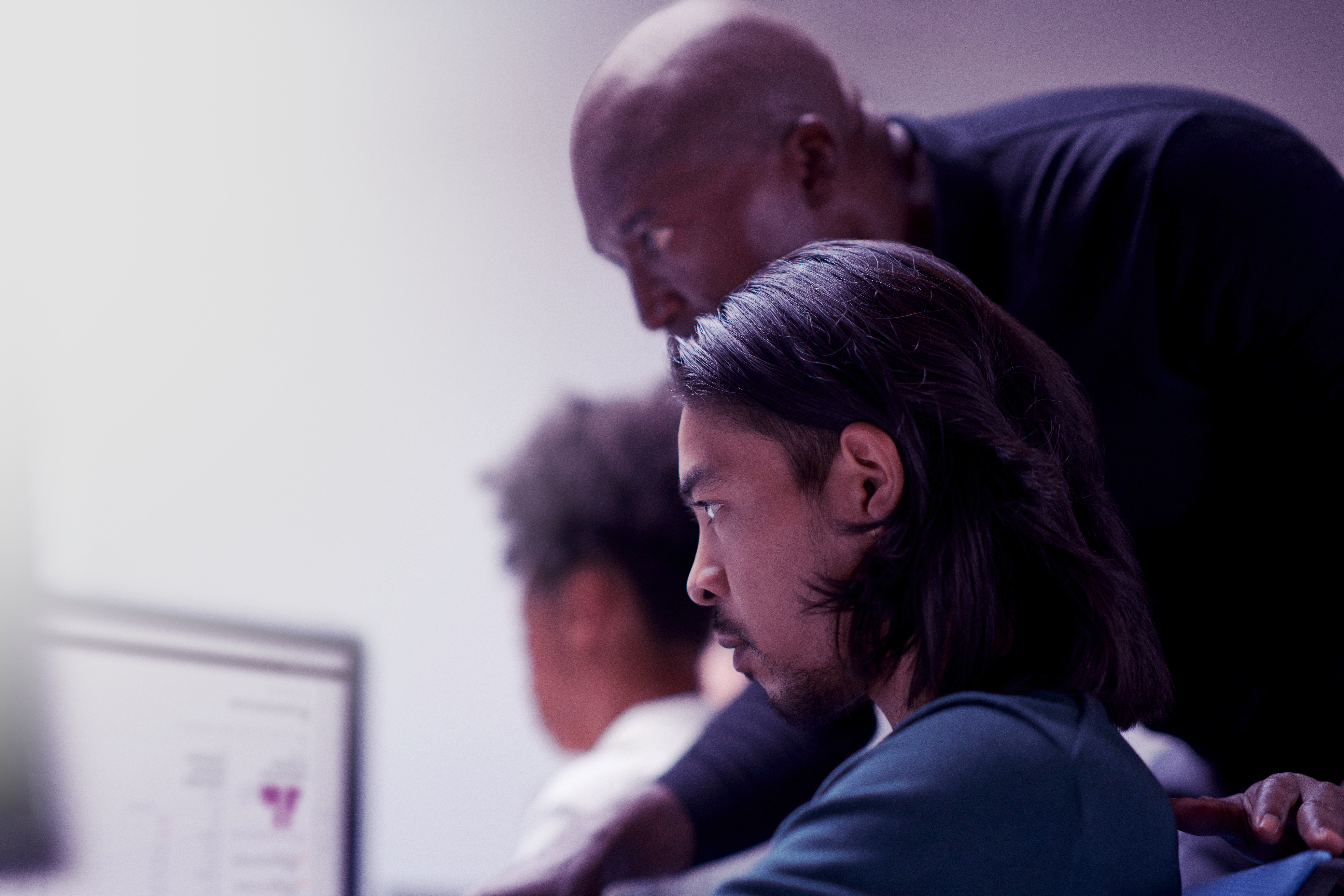 A mentor leans over to guide a focused learner who is working at a computer, with another person blurred in the background. The scene takes place in a classroom or training environment with soft, cool lighting.