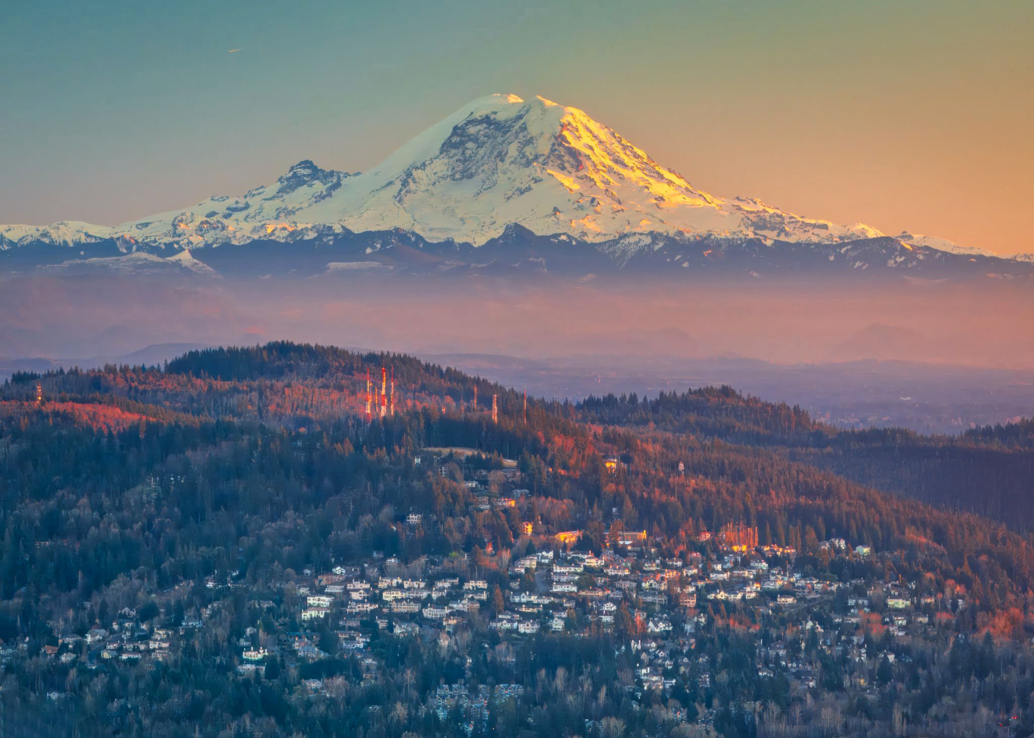Sunset light illuminating the snow-capped peak of Mount Rainier above rolling forested hills.
