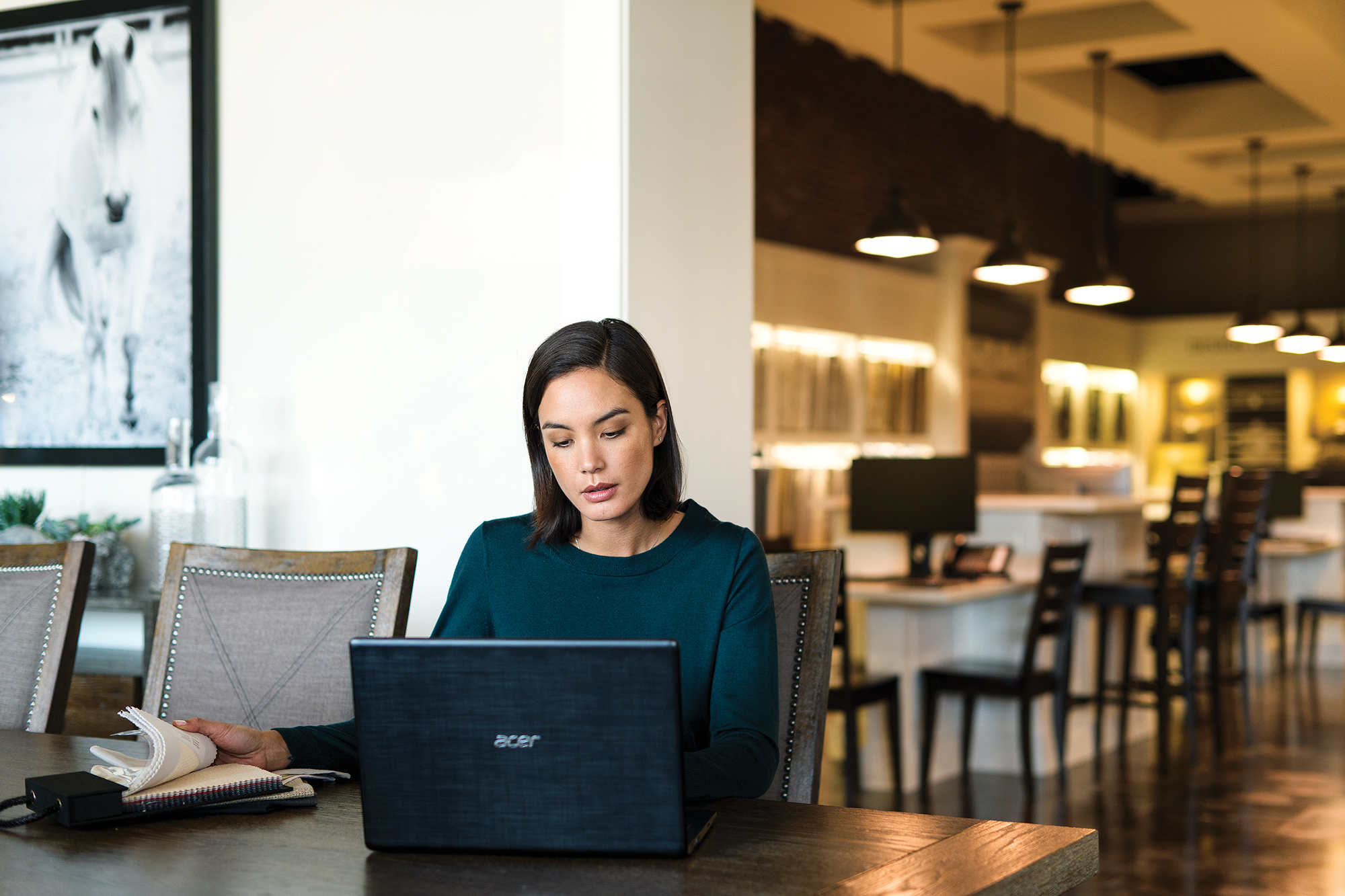 Female first line worker sitting at table in commercial retail store using Acer laptop. She is flipping through a book of fabric sample swatches, while looking at laptop