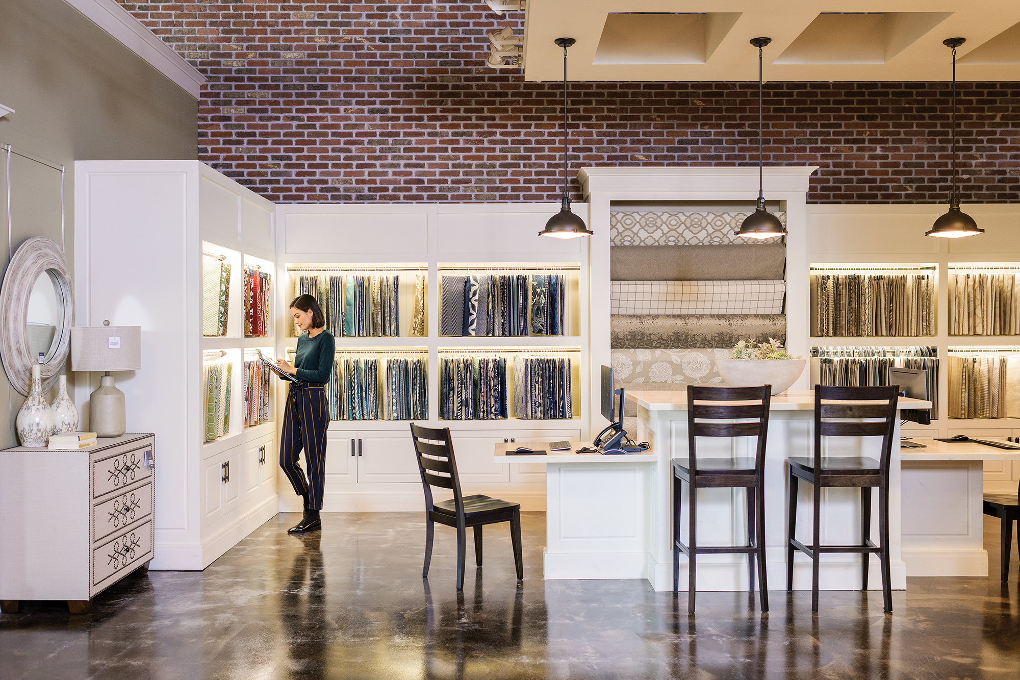 Female first line worker in retail store, standing directly in front of merchandising display wall filled with fabric panels while using pen on ASUS convertible laptop folded open as tablet (screen not shown)