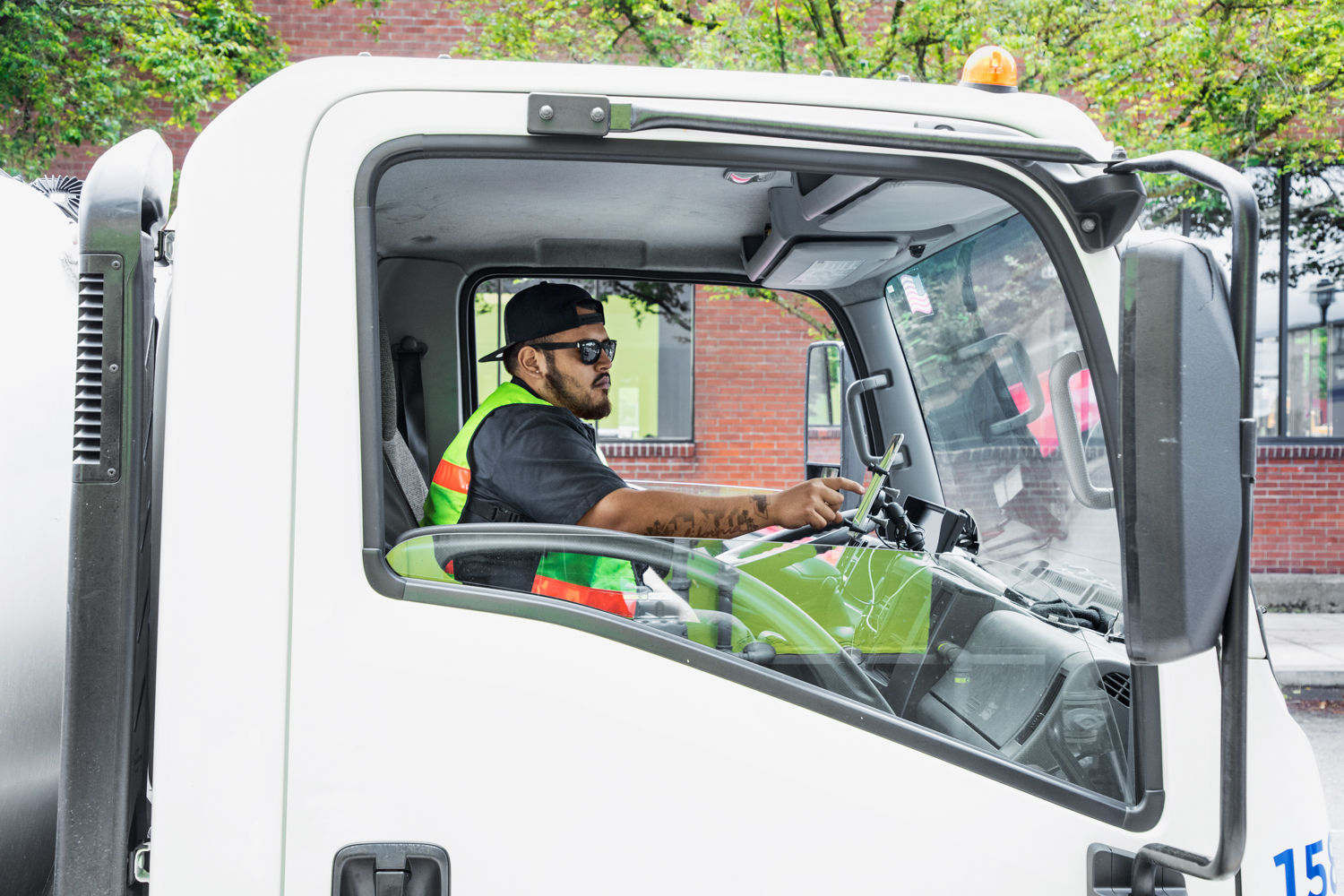 Male worker sitting in driver&lsquo;s seat of small loading truck using tablet mounted on dashboard. Photo taken from outside passenger door. Man&lsquo;s tattoos visible on arm