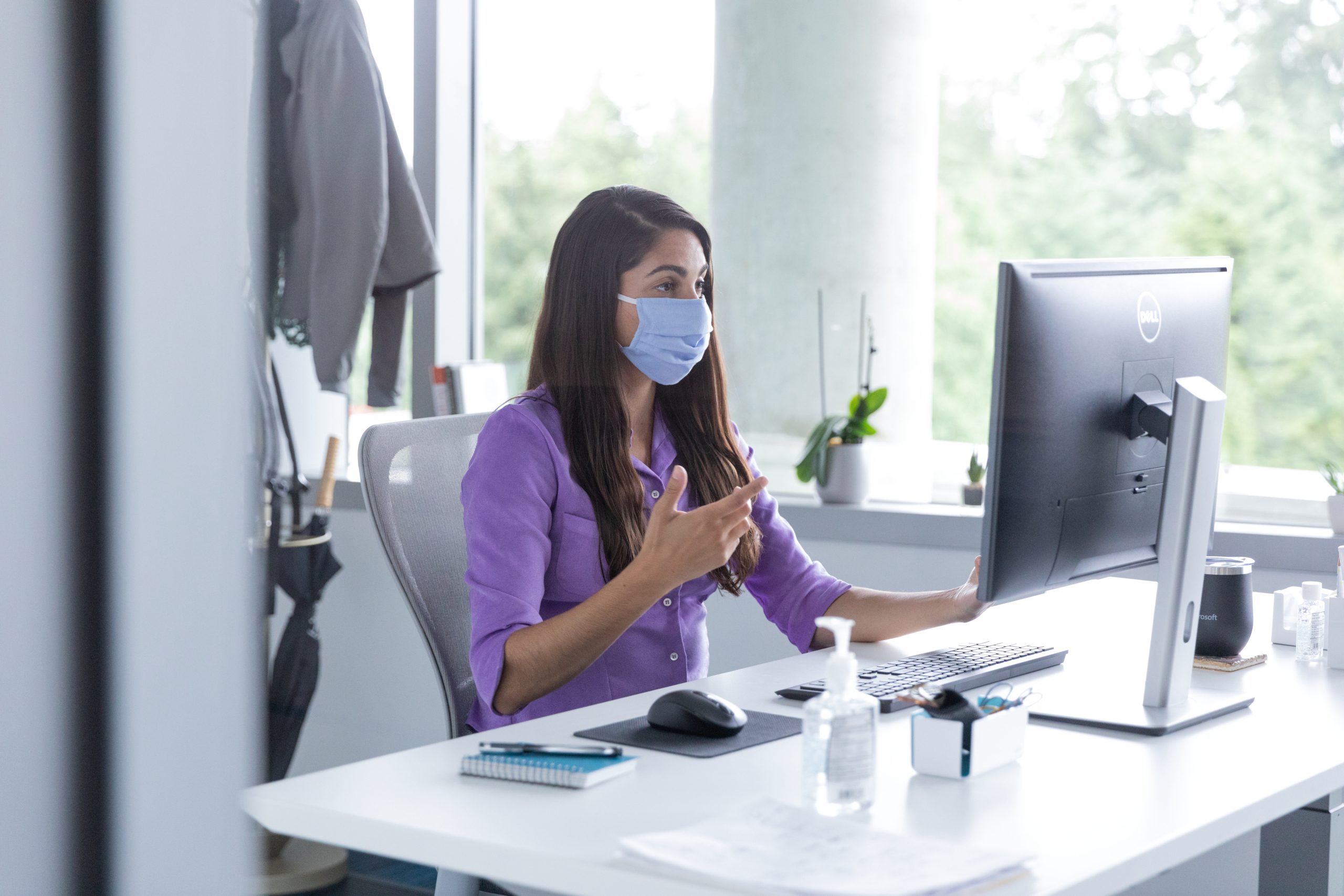 A female employee talking on a video call with hand sanitizer on her desk, socially distancing from others. Hybrid Workplace collection