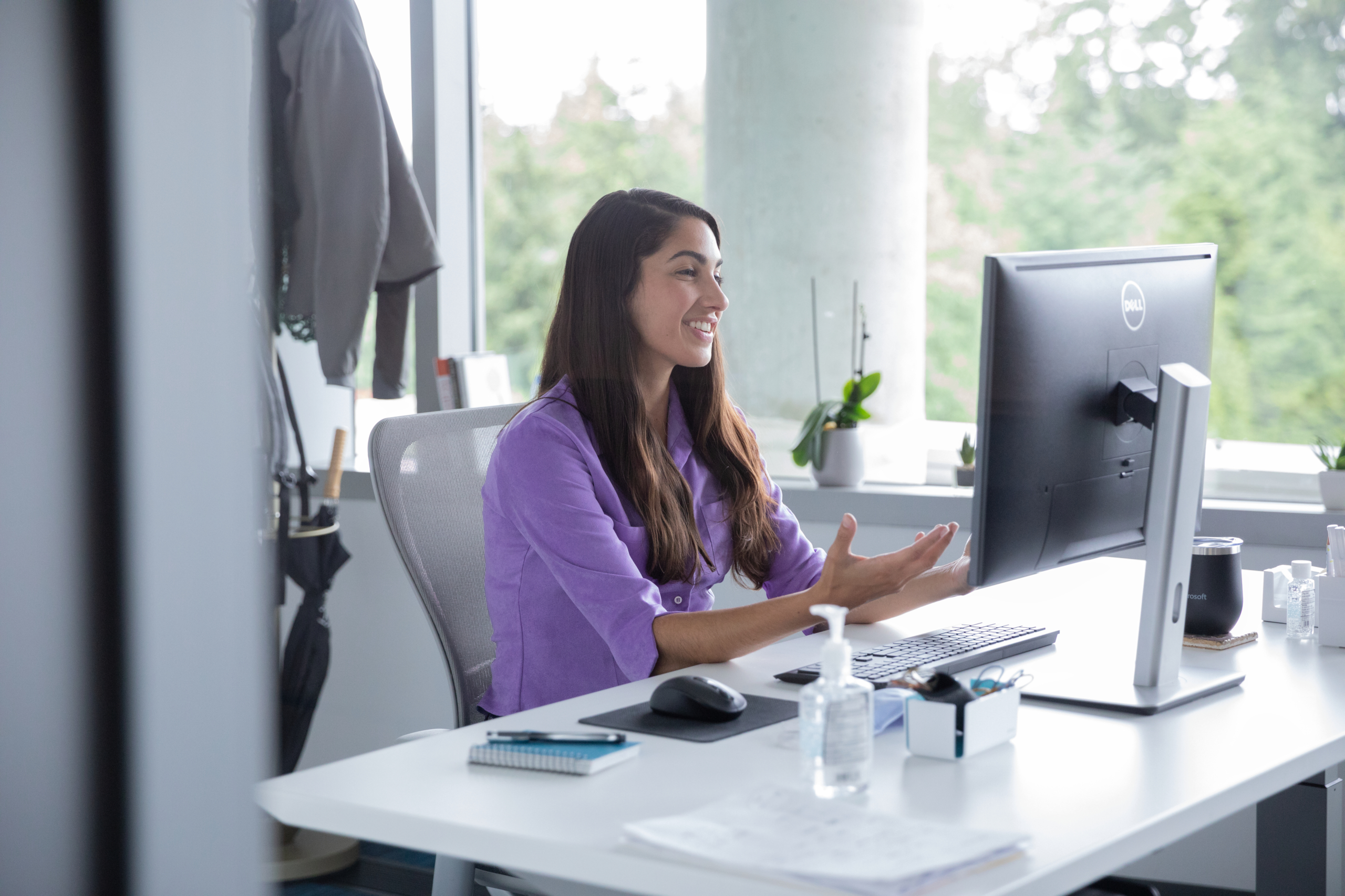 A female employee talking on a video call with hand sanitizer on her desk, socially distancing from others. Hybrid Workplace collection