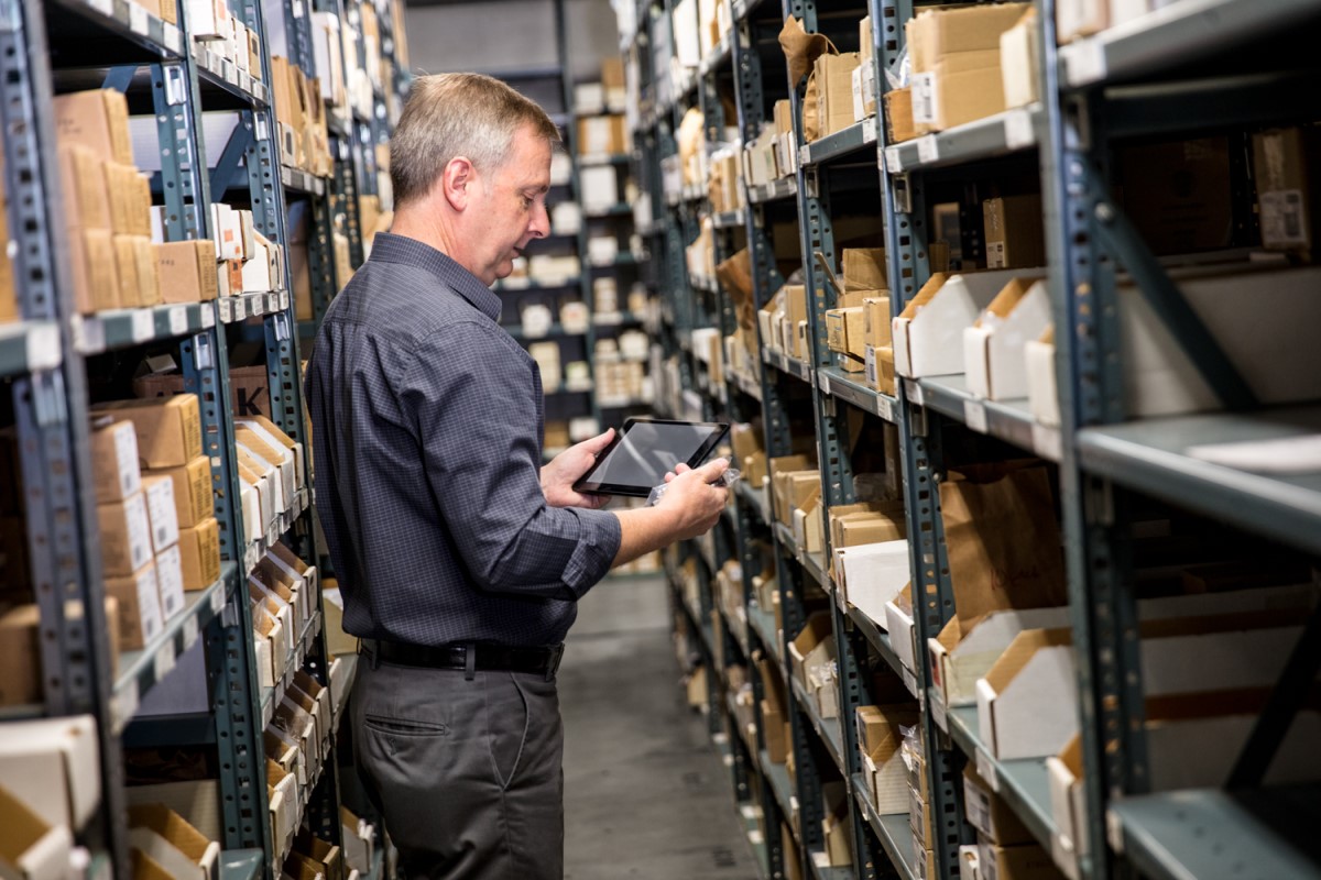 Male employee in warehouse storage room holding a tablet in one hand and small item in the other. He is standing between metal warehouse racks filled with pacakges