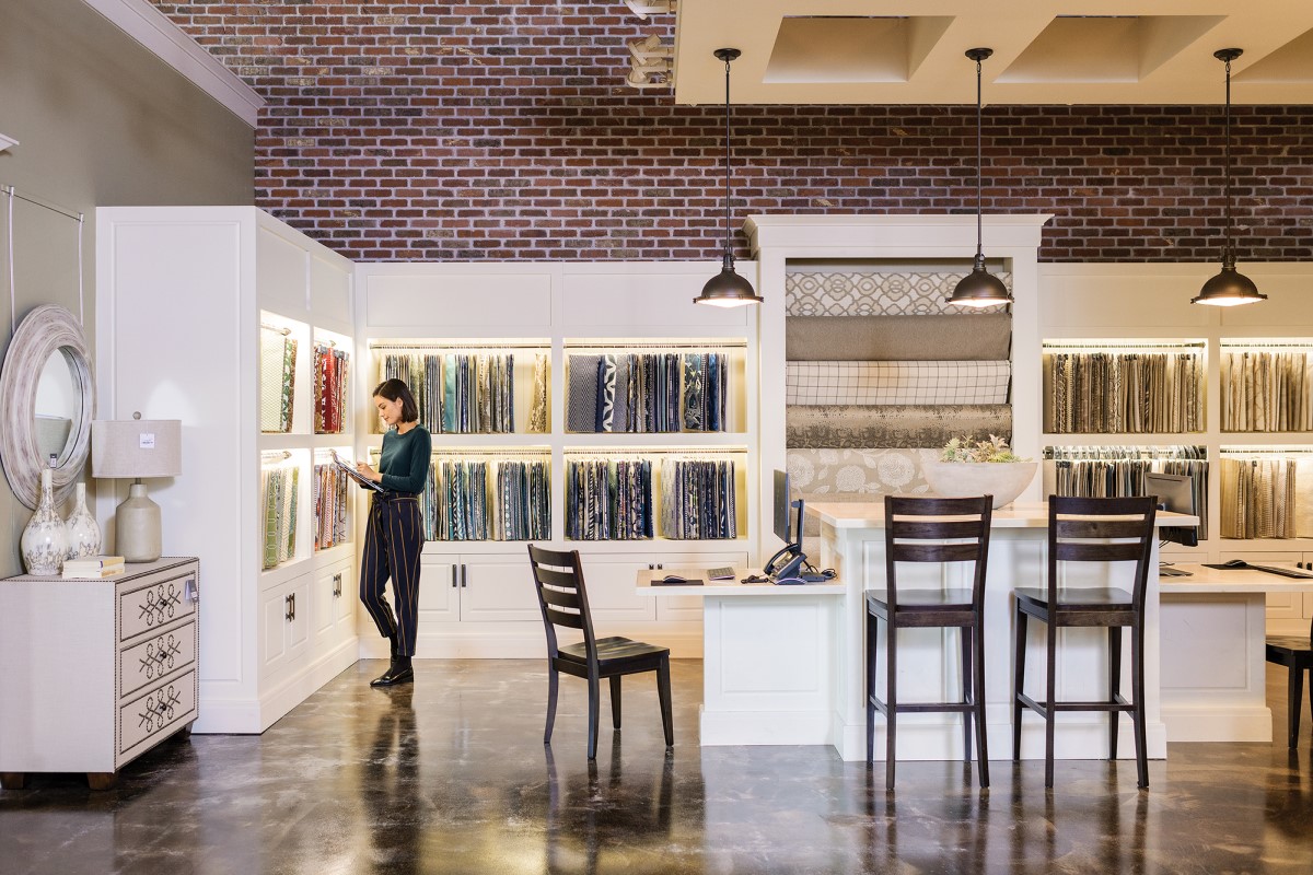 Female first line worker in retail store, standing directly in front of merchandising display wall filled with fabric panels while using pen on ASUS convertible laptop folded open as tablet (screen not shown)