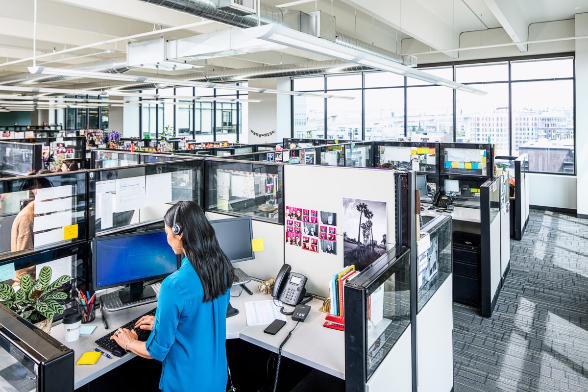 Female call center employee standing at cubicle desk, using desktop computer. Two monitors shown (one with Windows 7 log in screen visible) and a phone on desk. There are several other cubicles in the call center office
