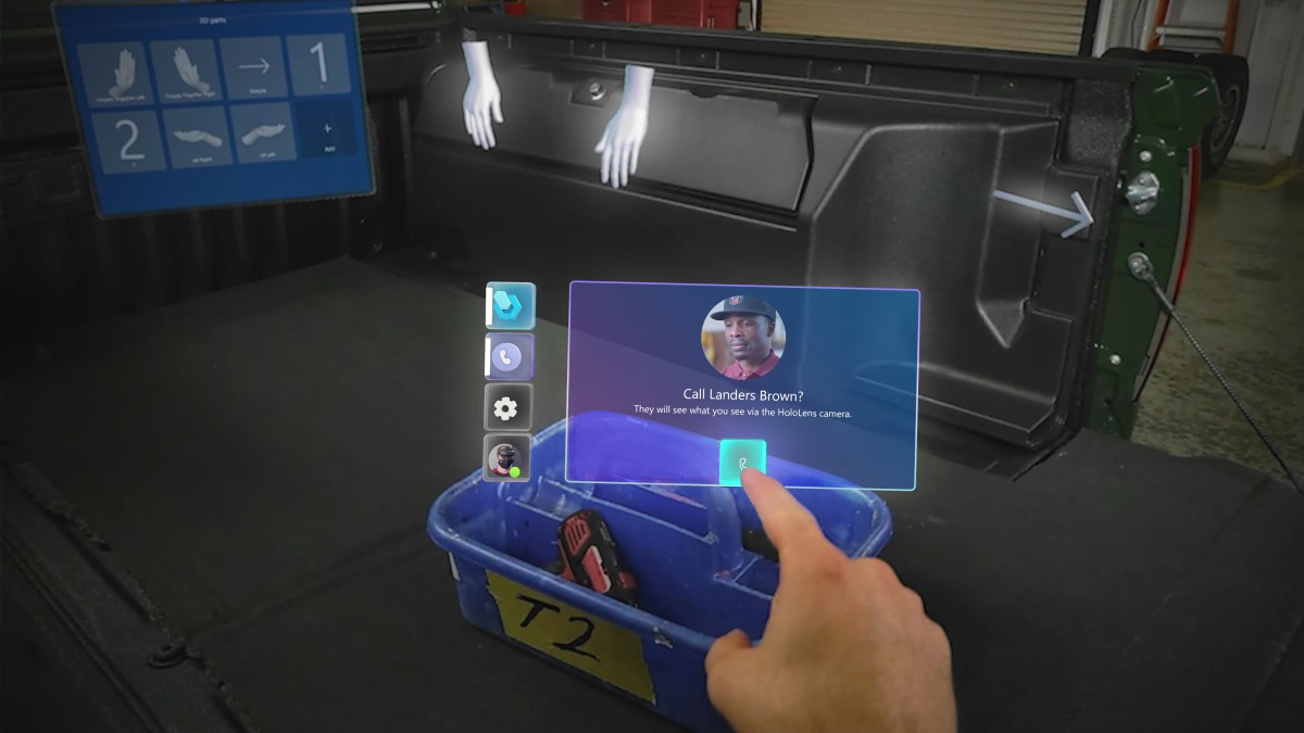 A user&rsquo;s mixed reality view from their HoloLens, showing their hand using touch controls for starting a holographic video call with a remote colleague