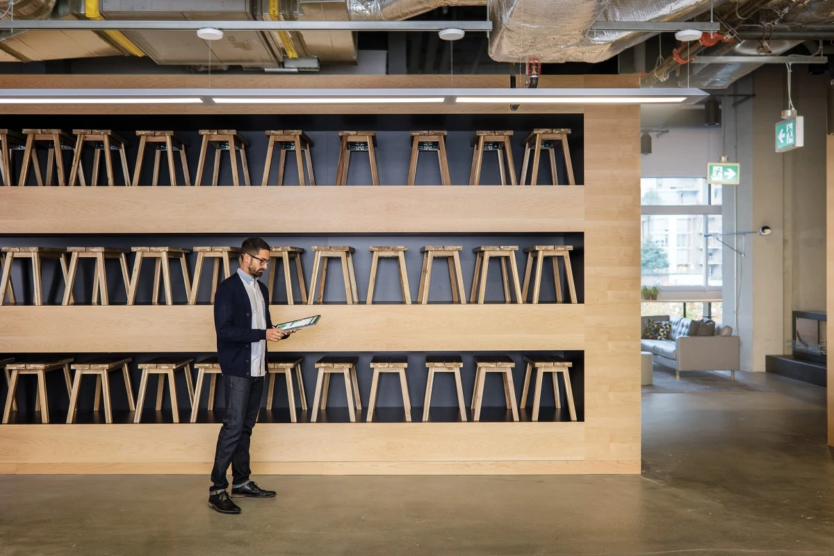 Male small business worker standing in front of three large shelves filled with dozens of wood stools. He is using an HP convertible laptop (opened as tablet) and pen. Screen is partially shown depicting bar charts and graphs
