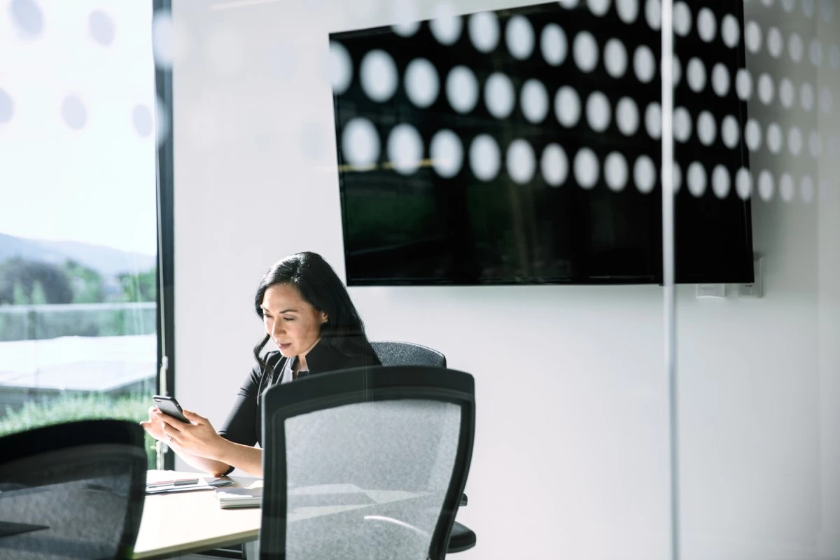 Female corporate office worker using phone at conference room table. There is a large monitor behind her (blank screen shown)