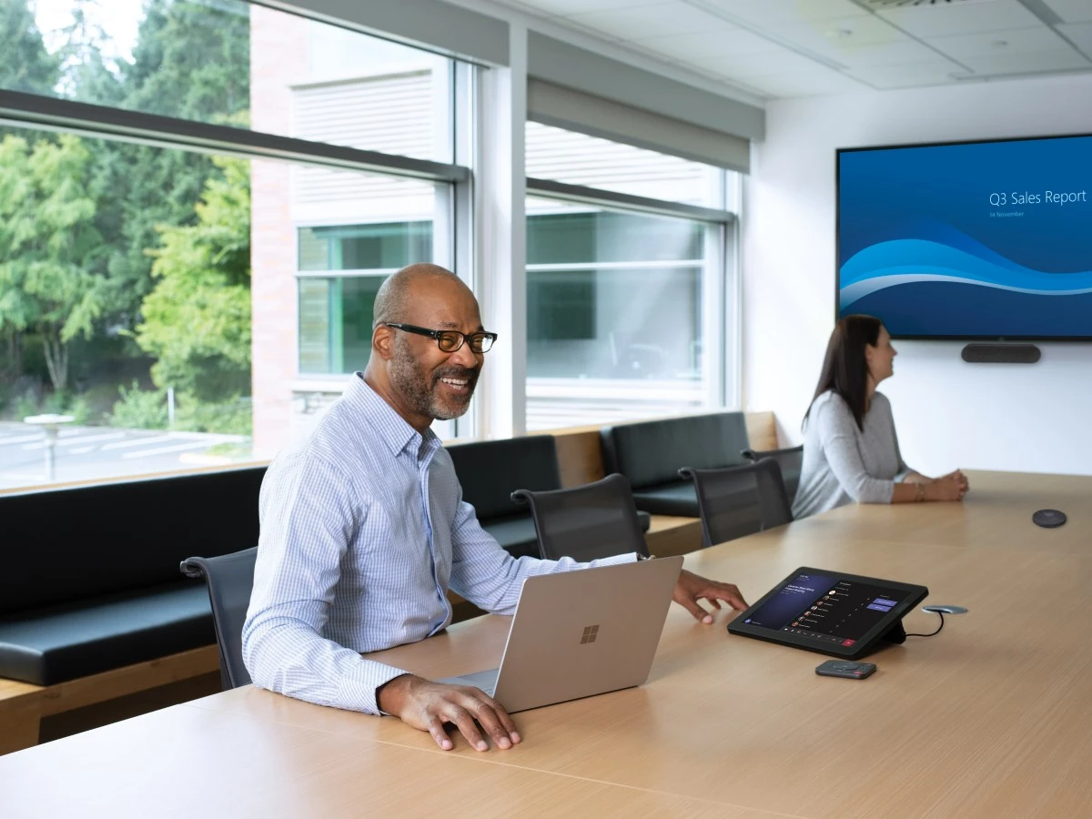 One male and one female in a large conference room featuring an HP Teams Meeting Rooms touch display joined to a Teams Meeting. Screensharing on a large mounted display and one Surface Devices in view