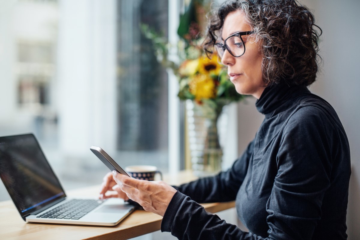 Mature businesswoman sitting at cafe looking at her cell phone while working on laptop computer