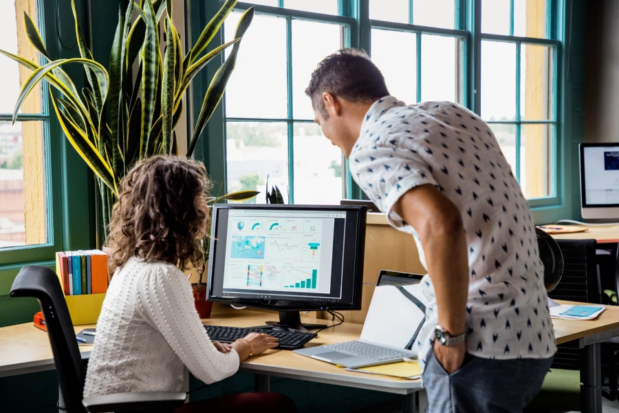 A woman sitting at a desk using a computer next to a man standing, looking at the screen