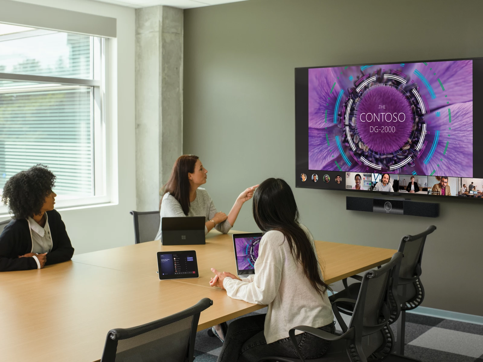 Three females in a large conference room featuring an Poly Teams Meeting Rooms touch display joined to a Teams Meeting. Screensharing and remote participants shown on a large mounted display and two Surface Devices in view