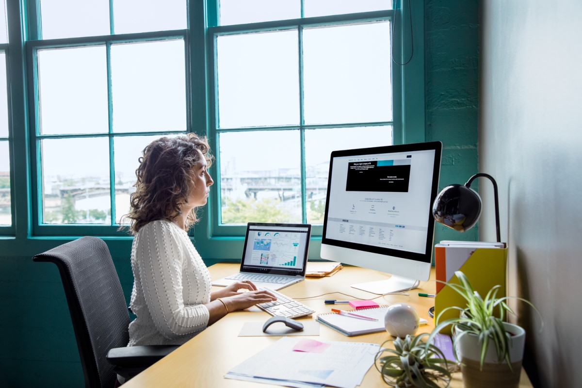 Female office worker sitting at desk using an all-in-one computer in casual office with a laptop also on desk