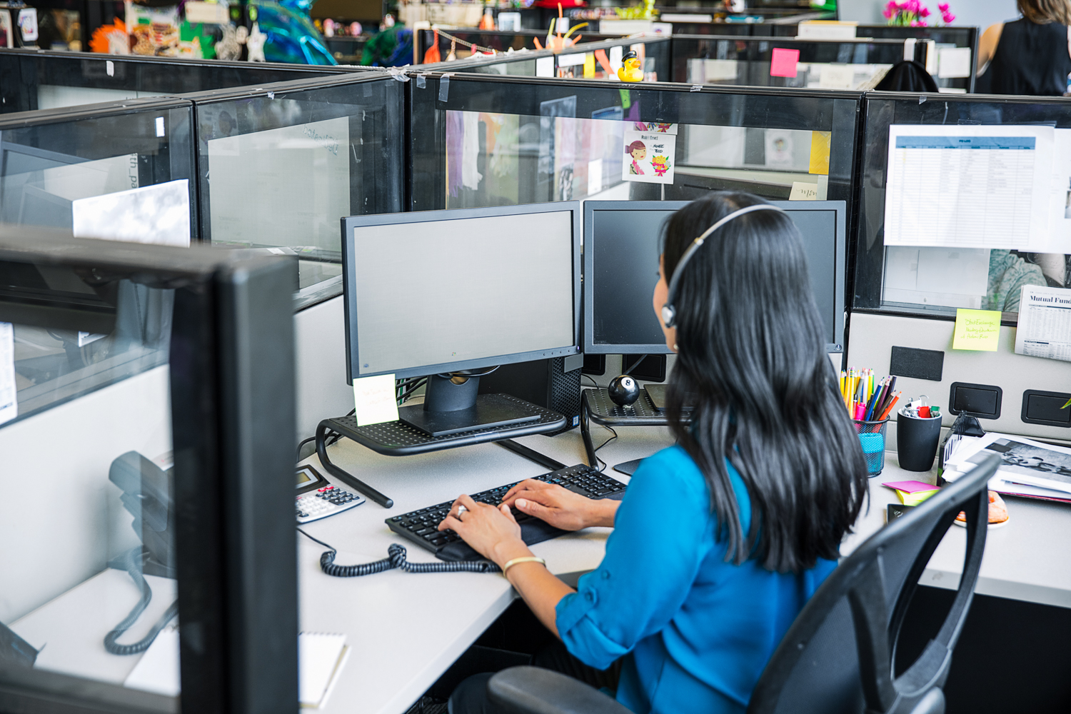 Female call center employee sitting at cubicle desk, using desktop computer with both hands on keyboard. Two monitors shown (no displays shown on screens)