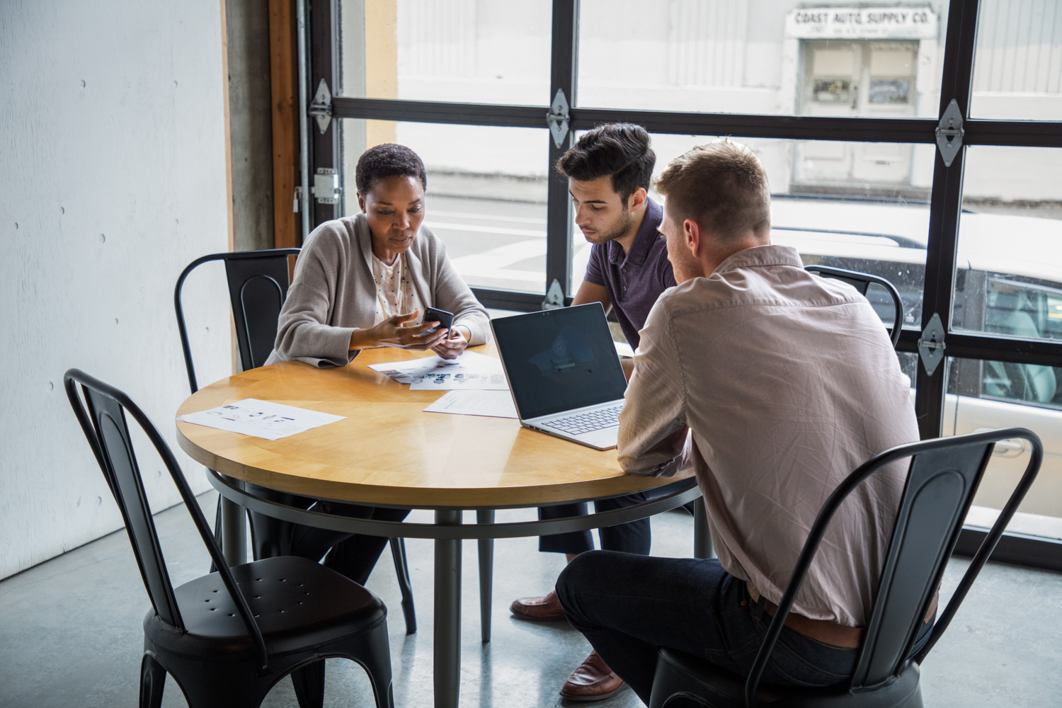 One female and two male workers sitting at round table in casual office setting or caf&eacute;/coffeehouse. The woman is holding a phone to show something to one of the men. The other man is using a Surface Laptop