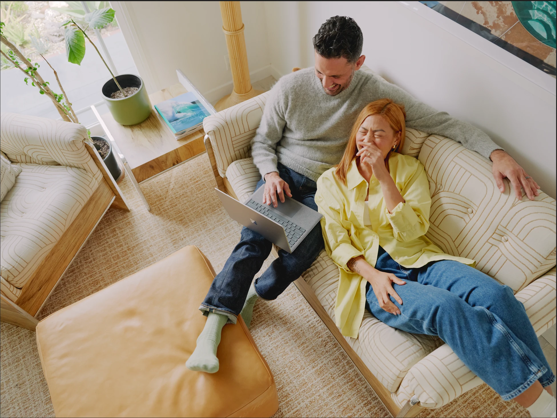Microsoft 365 lifestyle photography of a male and female adult on a couch smiling, laughing and looking at a Surface Laptop 5