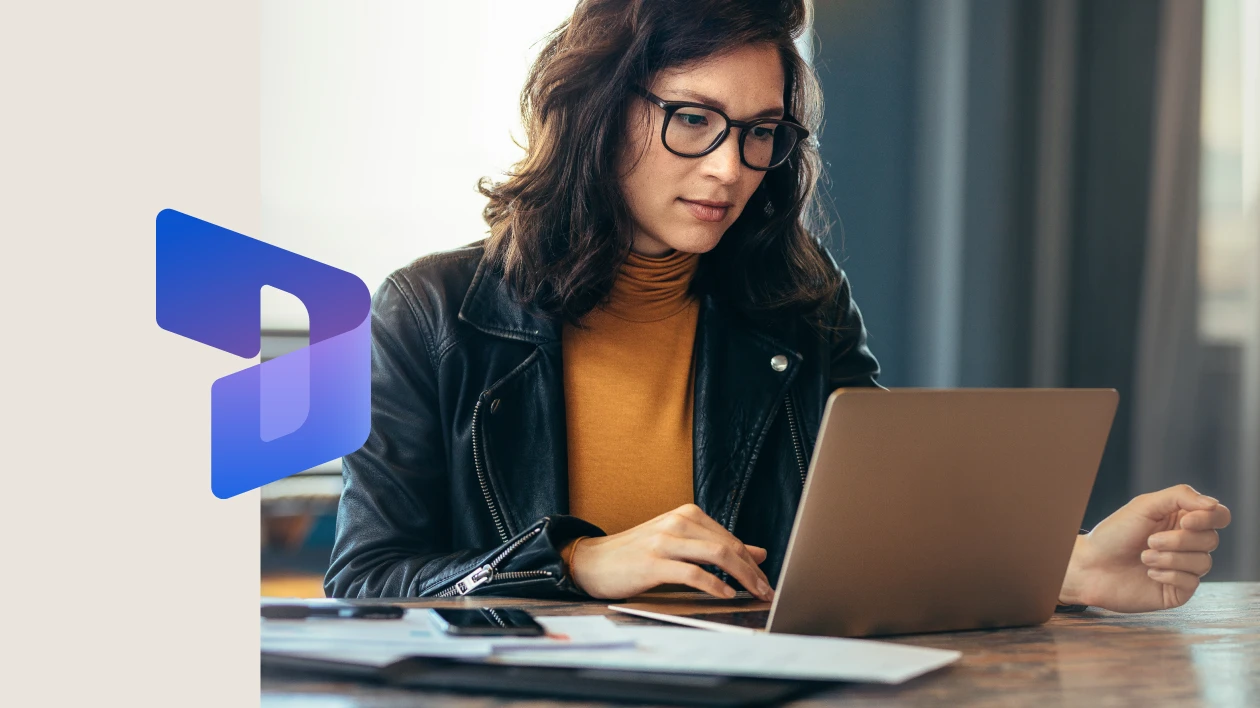 Businesswoman sitting at a table in an office, typing on her laptop with focus. The image is overlaid with the Dynamics 365 icon on the left