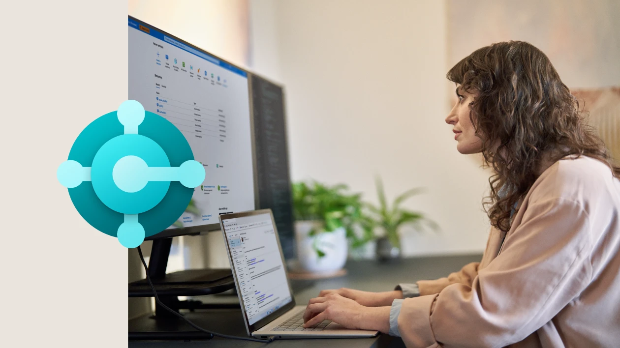 Side profile of a person typing on a laptop while looking at a large monitor screen behind it. The image is overlaid with the Business Applications icon on the left