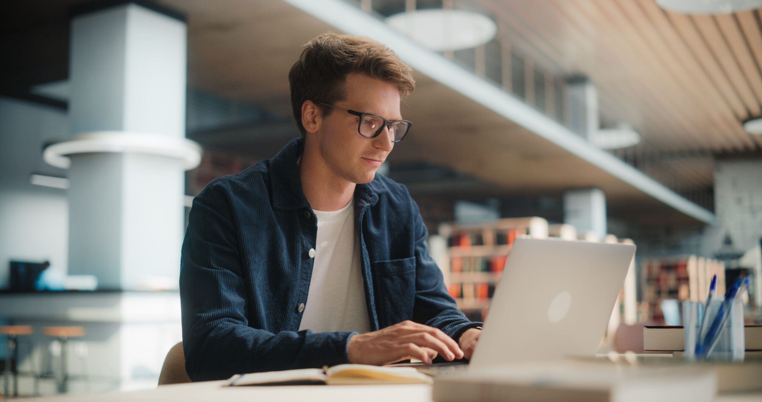 Concentrated Young Caucasian Male Student Working on His Laptop in a Modern Library. Man Engaged in Academic Research With Books Around, Focused on Screen