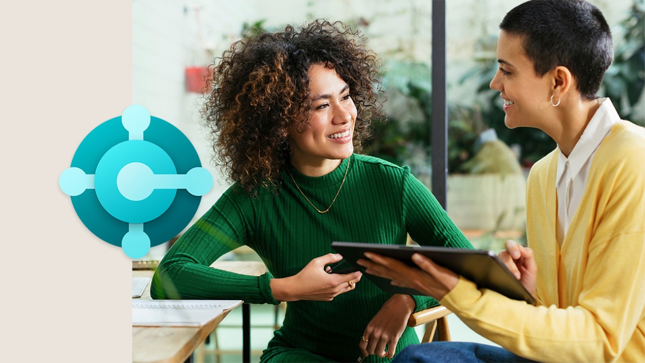 A woman in a green shirt talking to a woman in a yellow shirt whos holding a tablet. The Dynamics 365 Business Central icon is on the left side of the image
