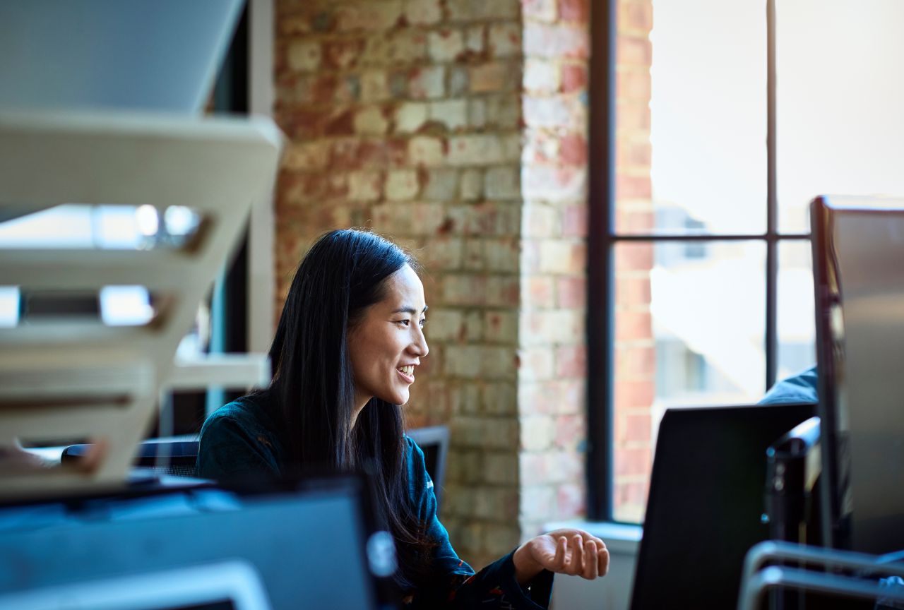 Office worker, smiling and looking away, in contemporary studio with bare brick walls, sitting in front of a desktop computer