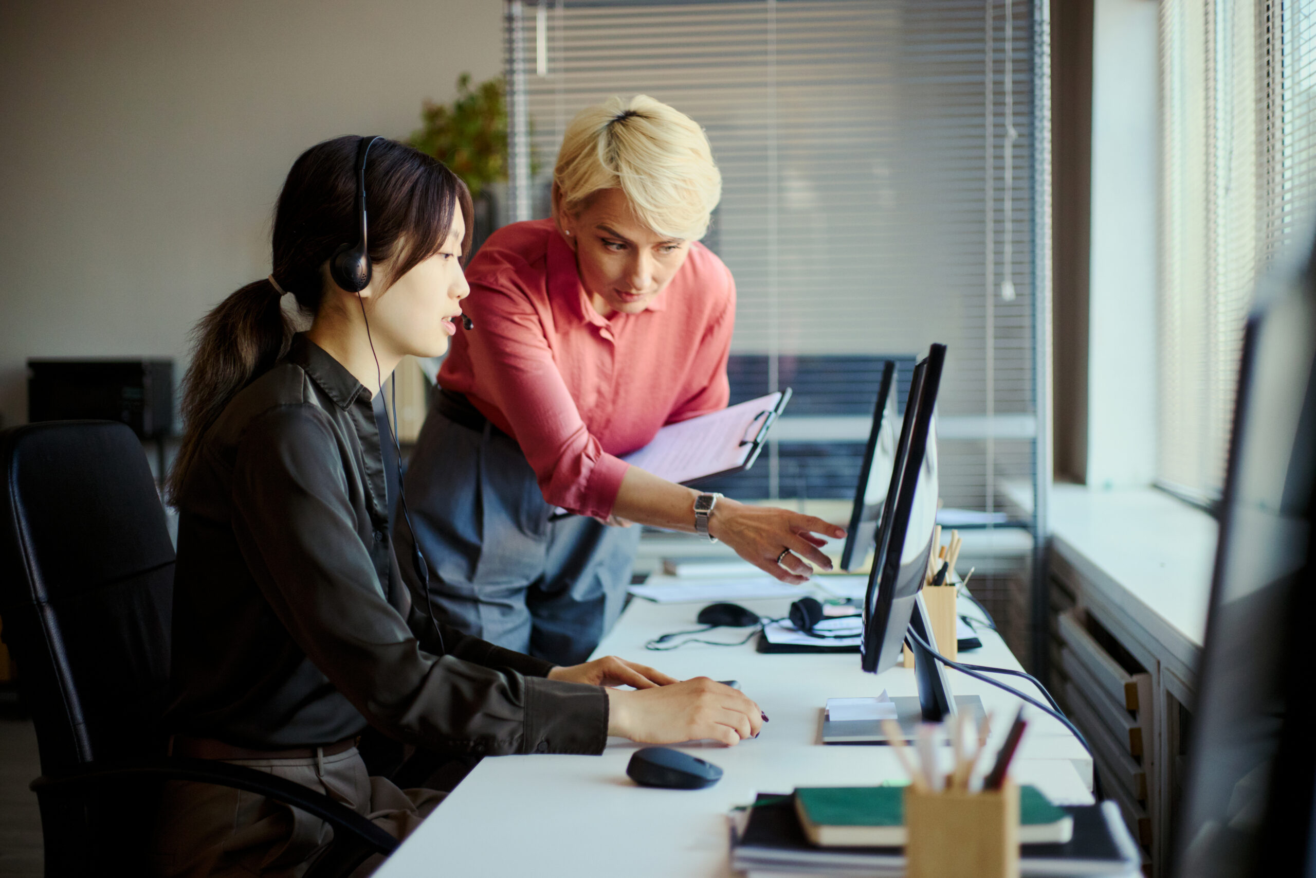 Young adult Asian woman working at call center desk while middle aged Caucasian woman guiding on computer, showing customer support teamwork. For service training, CRM, outsourcing marketing