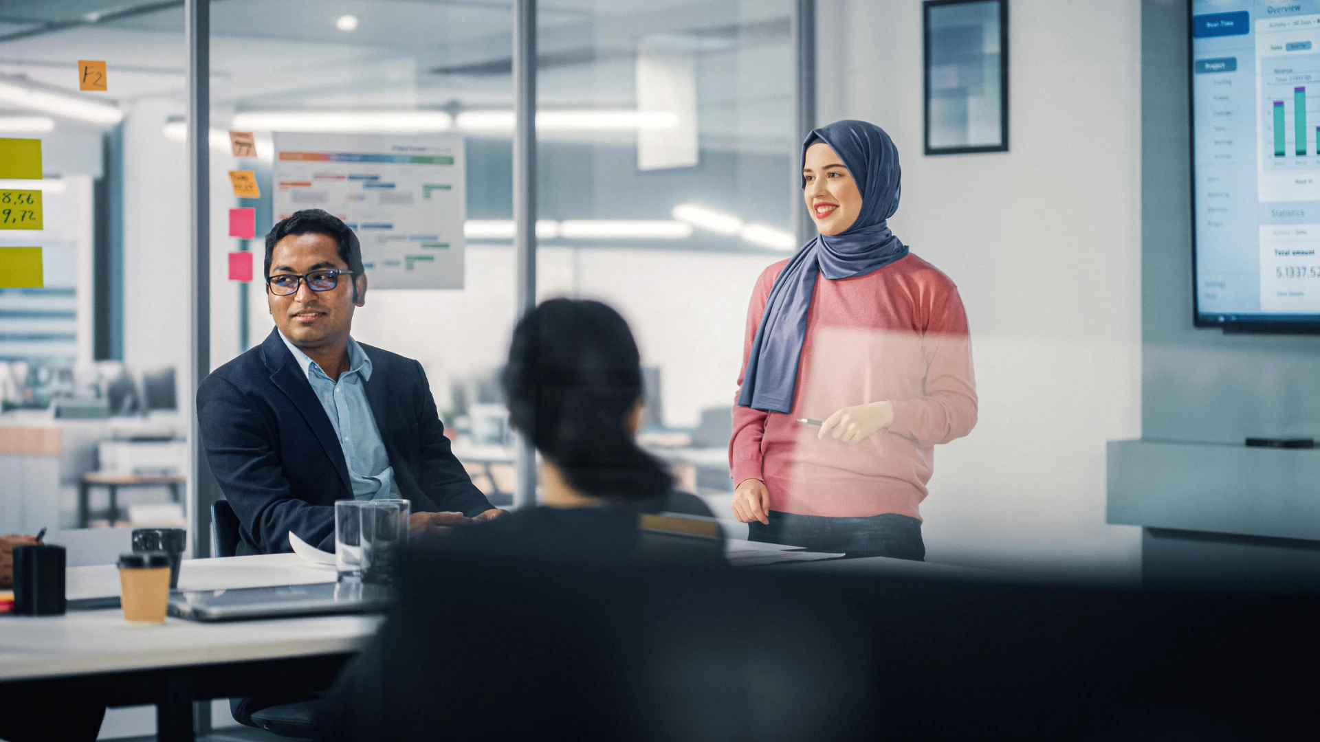 Colleagues collaborating in a modern office meeting, with one person presenting while others review project plans and data on a screen