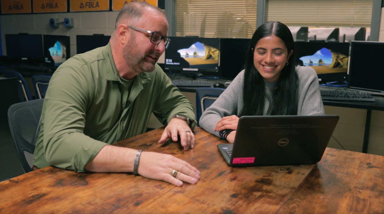 An educator and student sit together at a table in a classroom and look at the screen of a laptop