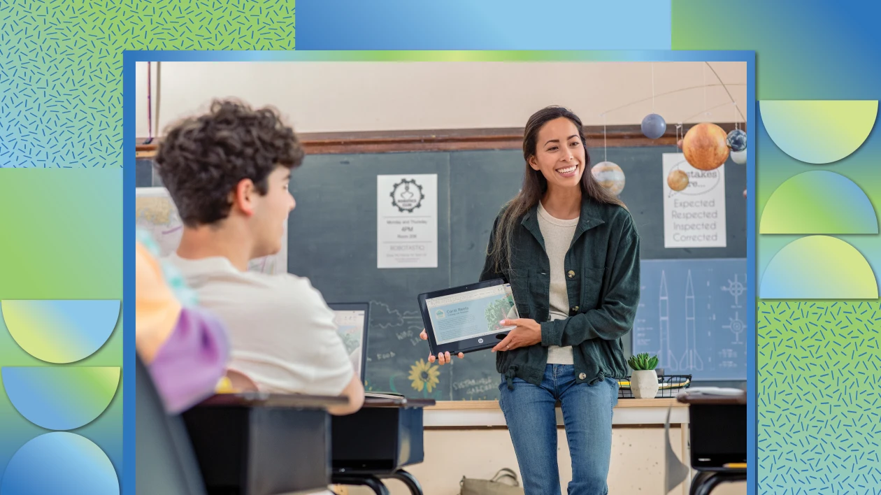 A teacher holding a folded laptop in front of students in a classroom