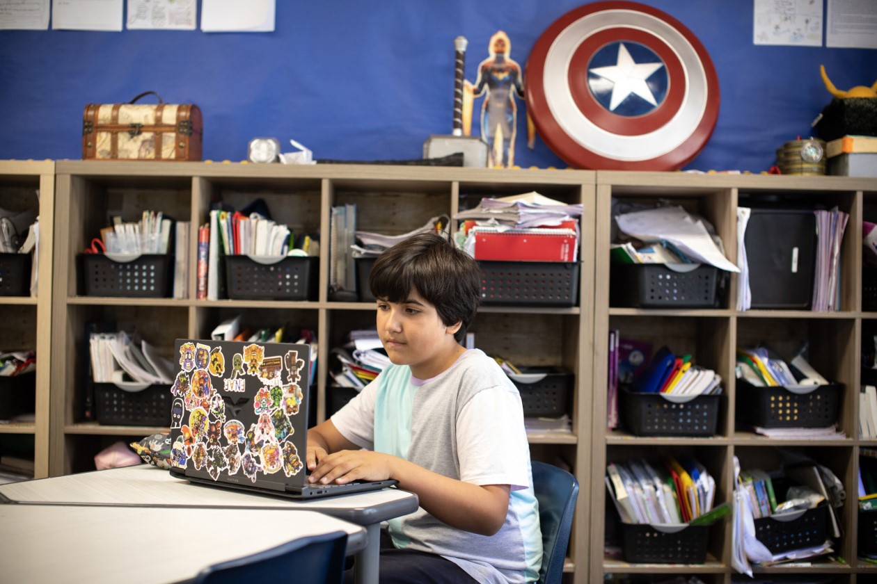 Decorative. A student sitting at a desk in a classroom and working on a laptop. There are bookshelves with bins of notebooks and folders behind the student