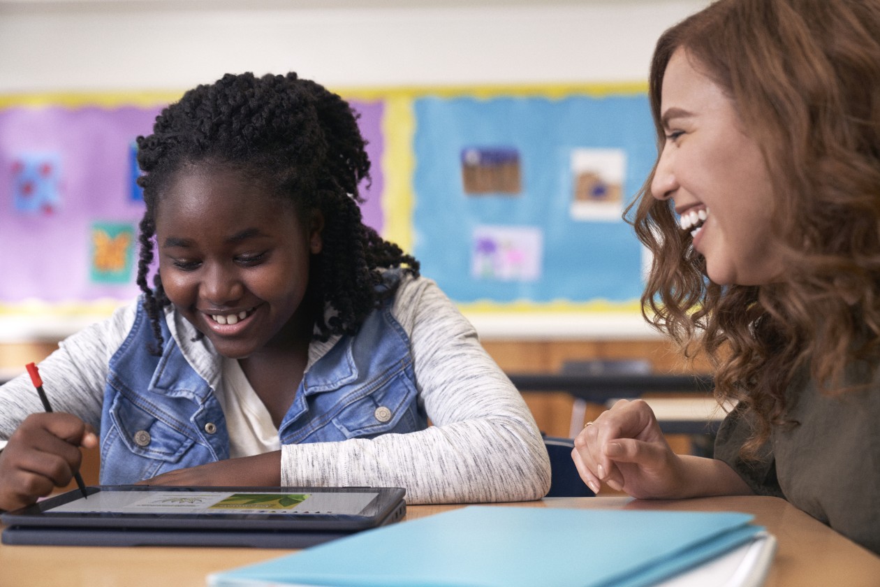 A student uses a digital pen to write on a laptop while an educator smiles encouragingly