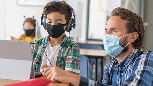 A child with a face mask and a man with a face mask and headphoneslooking at something on the Surface Pro.