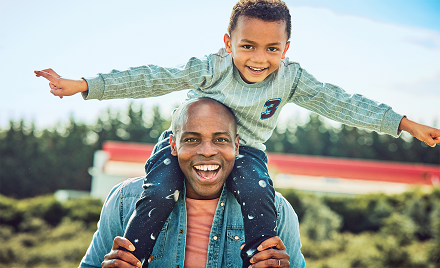 A man with a small boy on his shoulders walks outdoors. The two are smiling, and the child has his arms out like an airplane
