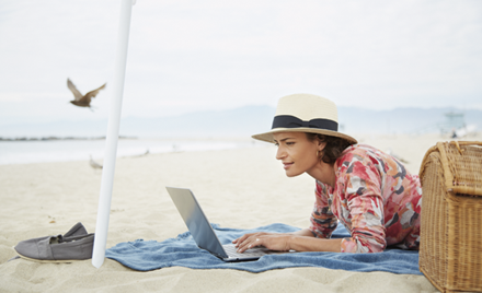A woman works remotely under a parasol, laptop open on a beach towel