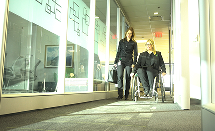 Image of two workers chatting in an accessible workplace hallway, one standing, the other from a wheelchair