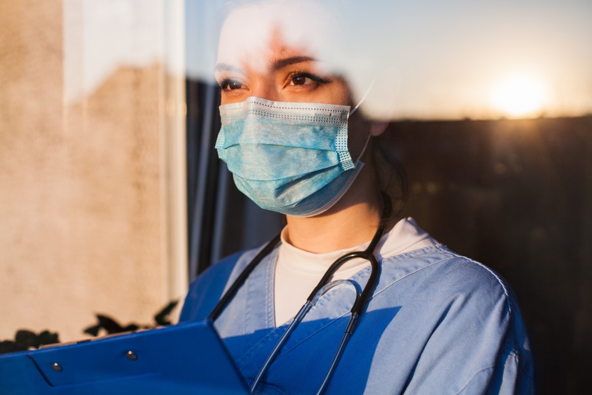A woman standing in a window, wearing nurses scrubs and a PPE mask., holding a clip board