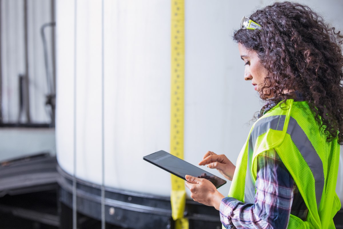 Female worker wearing neon vest and safety glasses using tablet. Industrial vats visible in background