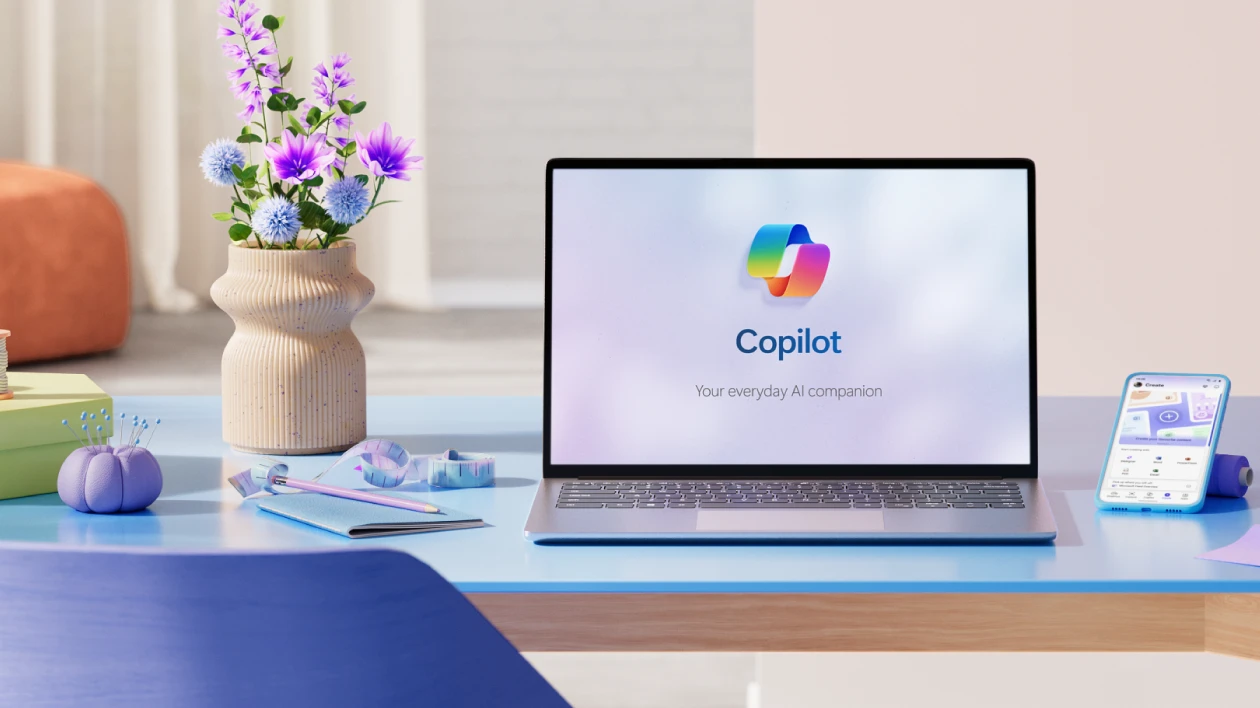 A laptop with the Copilot icon on screen on a blue desk with a vase of purple flowers, sewing cushion, and cell phone