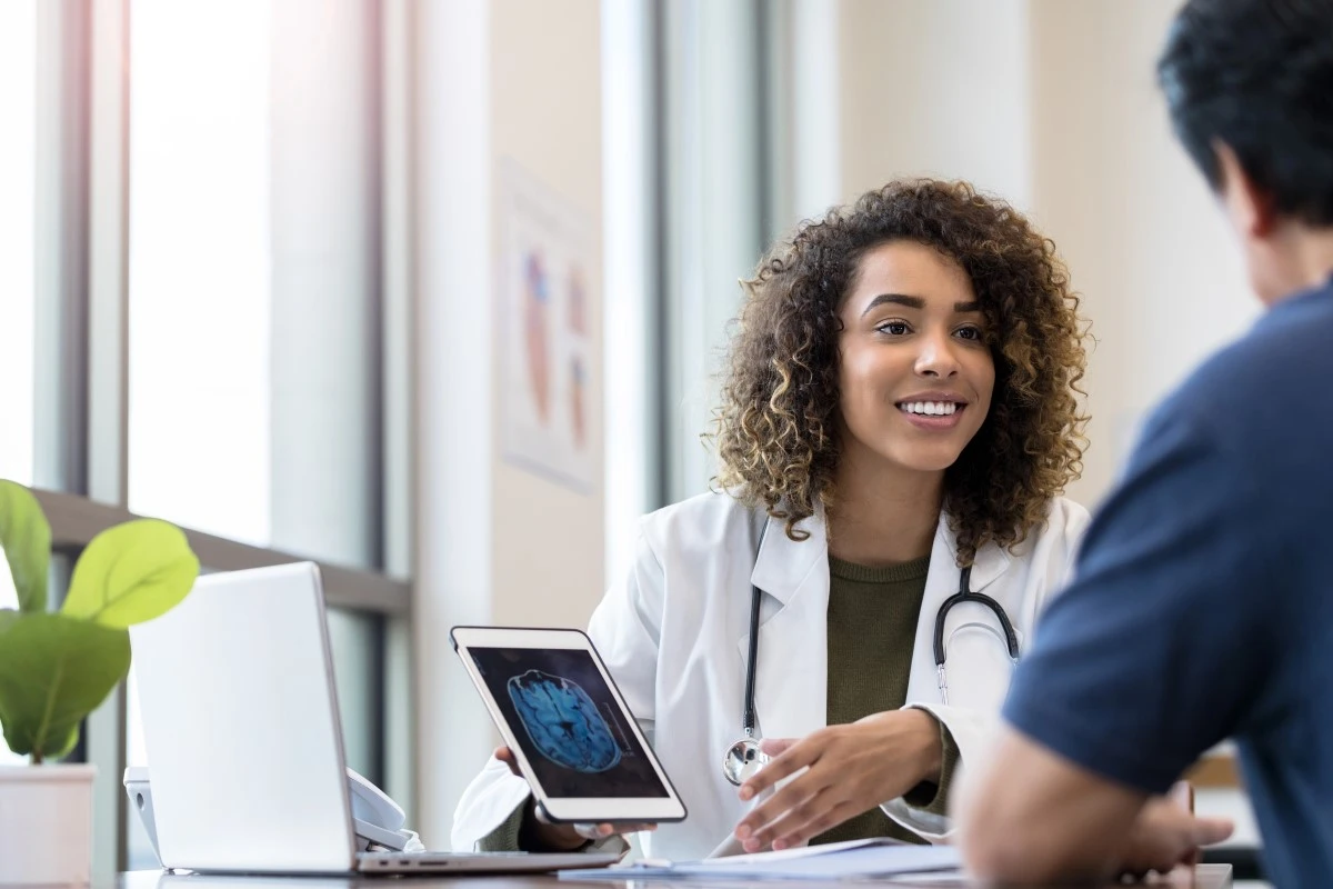 Female clinician w/ brown skin and brown shoulder length curly hair shows computer screen with unclear image of results to patient