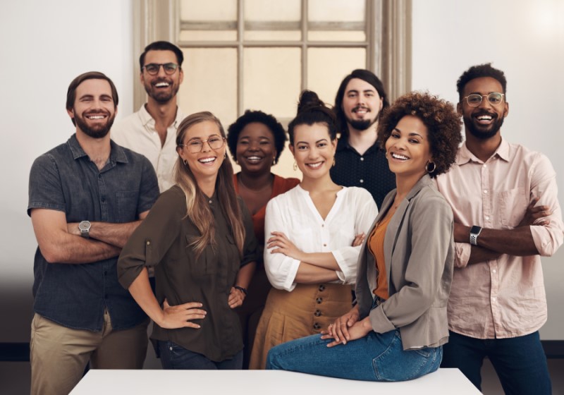 8 professionals of varied ethnic and age backgrounds in casual attire grouped together, smiling at the end of a conference room