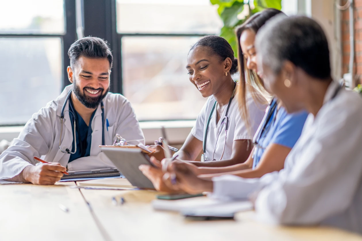 A small group of four medical professionals sit around a conference table as they meet to discuss patient cases. They are each dressed professionally in scrubs and lab coats as they focus on working together