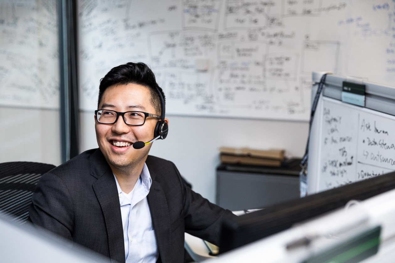 Male worker sitting at cubicle desk in financial office, wearing glasses and phone headset. He is smiling and looking up to his right. A large whiteboard covered in black written text is on wall behind him. A smaller whiteboard also partially visible on his desks cubicle wall